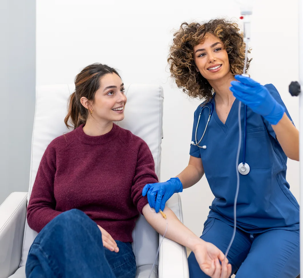 A nurse adjusting an IV line for a smiling patient in an infusion chair